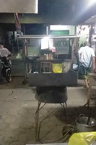 The video depicts a dimly lit outdoor scene, possibly a street or alleyway, at night. A person in a white shirt and patterned garment stands near a food stall. The stall is illuminated by a bright light and displays various items, including bottles and bags. A large, cylindrical cooking vessel sits prominently in the foreground. Several motorcycles are parked nearby. Underneath a colorful umbrella, a group of people are gathered, some of whom appear to be interacting. The ground is paved with interlocking stones. The overall atmosphere suggests a casual gathering or a busy street food vendor operation.