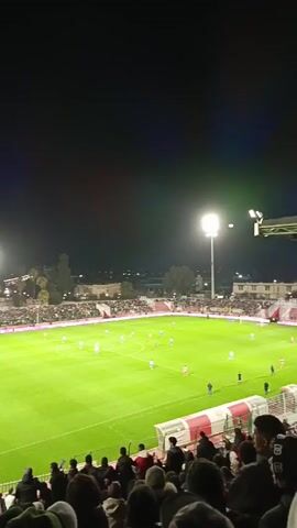 The video shows a soccer game at night. The stadium is packed with fans, and the field is lit up by bright floodlights. The camera pans across the field, showing the players and the stands. Someone raises their fist in the air. The fans are chanting and cheering. Buildings are visible outside of the stadium.