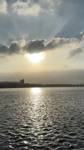 The video shows a wide shot of a body of water with ripples. The sun is partially obscured by clouds, casting rays of light. On the horizon, there is a silhouette of a city skyline with buildings and a bridge. The water reflects the sunlight. The sky is cloudy with some areas of brightness. The overall scene appears to be a landscape during daylight hours.
