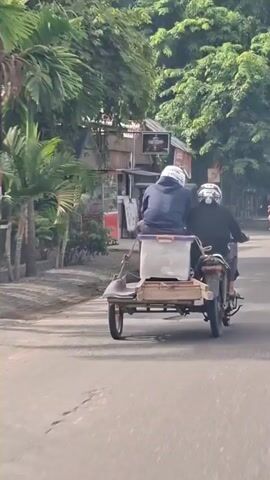 The video shows a motorcycle with a sidecar driving down a road. Two people are riding in the sidecar, one in front and one behind. Both individuals are wearing helmets. The sidecar has a large plastic container on it. The road is paved and appears to be in a rural or suburban area, with trees and buildings lining the sides. The weather appears to be sunny.
