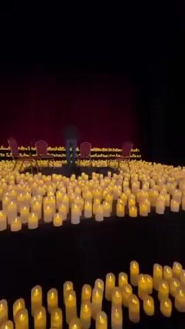 A person walks across a stage covered with hundreds of illuminated white candles. Four chairs are set up on a raised platform in the background, in front of red curtains.  A person holds a phone in the foreground to record the scene.