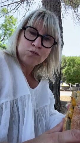 This close-up, selfie-style video shows a woman with blonde hair and black-rimmed glasses eating a sandwich outdoors. She is wearing a white blouse with ruffled sleeves. The background shows a tree trunk to her right and foliage and a clear sky to her left, suggesting a sunny day, likely daytime. The woman is holding a long sandwich with her hands and taking a bite. Her expression is one of enjoyment or concentration as she eats. The setting appears to be a park or natural area. There is no visible text in the video.
