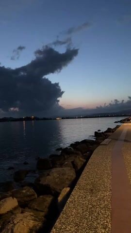 The video shows a tranquil view of a body of water at dusk. Dark, ominous clouds dominate the sky, with hints of pink and blue from the setting sun peeking through. Lights from a settlement across the water are reflected in the calm surface, offering a glimpse of civilization. The foreground consists of large rocks lining the water's edge, next to a paved walkway. The overall atmosphere is serene, tinged with the potential for an impending storm.