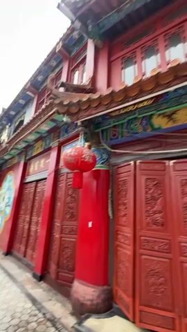 A video captures the side of a red building with ornate Chinese architecture. Red double doors with intricate carvings line the wall, and a large red lantern hangs from a column. A circular mural featuring a lotus flower and what appears to be a dragon or mythical creature adorns the wall between the doors. The building's upper level features windows and decorative colorful trim. A cobblestone walkway runs along the front of the building, with a small white line painted on it. In the distance, trees and a car are visible. Towards the end of the video, two people on a white electric scooter ride past the building from right to left.