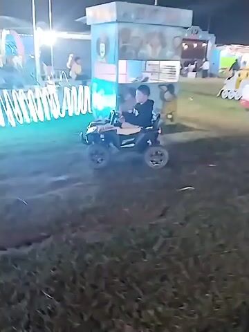 The video shows a night-time scene at what appears to be a fair or carnival. A small child is riding an electric toy ATV on a grassy area. There's a carousel illuminated with lights nearby, and several other structures that look like game booths or vendors, also lit up. The camera is shaky and the footage is a bit dark. In a few shots, a person with short black hair is partially visible in the foreground.