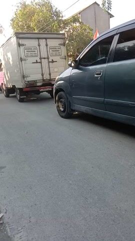 The video shows a street scene. A dark-colored SUV is parked on the right side of the road. To the left of the SUV, a white truck with a cargo container is visible. Several motorcycles are parked on the side of the road. One motorcycle is red and is occupied by a person wearing a light green jacket, black pants, a baseball cap, and a face mask. The person is leaning on the handlebars. Another motorcycle is black and is partially visible. In the background, there are houses and trees. The sky is clear and blue. The road is paved. The video appears to be filmed from a moving vehicle, possibly a motorcycle, as indicated by the slight shake and the angle of the shot. The orientation of the video is sideways, suggesting it was filmed with a phone held vertically.