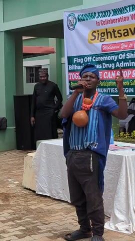 A man in traditional attire, including a blue and striped tunic, dark pants, and sandals, stands center frame speaking into a microphone. He has a gourd around his neck. He gestures with his right hand, which is raised with his index finger extended. Behind him, a banner reads "KADUNA STATE MINISTRY OF HEALTH In Collaboration With SIGHTSavers" and "Flag Off Ceremony Of Schistosomiasis MIDA Drug Administration. STOP SCHISTOSOMIASIS BEGIN WITH PREVENTION". To the left of the man, a speaker is visible. In the background, a man in a black tunic and cap stands near a green building. Several people in white hijabs and one person in a red hijab are seated to the left, some looking at a tablet. Other individuals are seated under a white tent in the background. The ground is paved with brick-like stones, and there is a small table covered with a white cloth to the right of the main speaker. The environment appears to be an outdoor gathering, possibly a public health event.