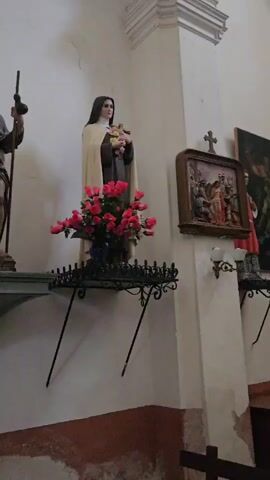 The video shows the interior of a church. A statue of a male figure holding a staff and a dog stands on a shelf to the left. Next to it is a statue of a female figure in religious attire holding a bouquet of flowers. A decorative shelf with wrought iron accents holds the flowers. To the right, a framed artwork depicts a religious scene with multiple figures. Below this artwork are two wall-mounted light fixtures. In the foreground, a tall, dark, spiral-shaped column is visible on the left, and a black portable heater with a reflective top stands near the center. The walls are white with some areas showing a reddish-brown discoloration at the bottom.