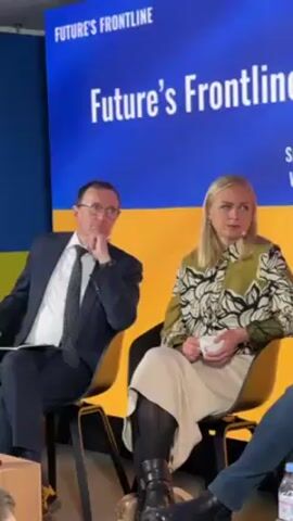 The video shows two people seated on a stage during a conference. The stage backdrop is a blue and yellow sign that reads "Future's Frontline." A man wearing glasses, a dark suit, and a tie sits with his chin resting on his hand. A woman in a floral print blouse and tan skirt is seated next to him, holding a white mug. The video focuses on the two individuals, and a small portion of a person wearing blue jeans is visible on the right side of the frame. The setting appears to be a conference or panel discussion.