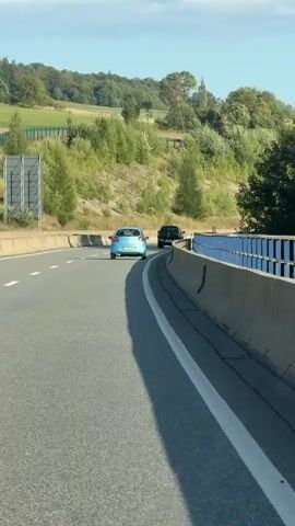 The video shows a blue car driving on a highway. The car is in the right lane and is following a black vehicle. The highway has a concrete barrier on the right side and a grassy embankment on the left. The sky is clear and blue. The sun is shining. The video is taken from another vehicle that is also on the highway.