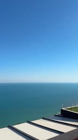A vertical video shows a wide expanse of turquoise water meeting a clear blue sky at the horizon. Several small, dark objects, possibly birds or drones, are visible in the sky. In the foreground, the edge of a rooftop with white awnings and dark trim is visible. The sun is shining, casting light on the scene.