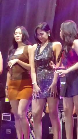 Three young women stand on a stage with a dark backdrop and purple lighting. The woman on the left has long dark hair and wears a brown top and an orange skirt. The woman in the center has dark hair, a sparkly black dress, and is dancing. The woman on the right has long dark hair and wears a red top and a black skirt. There are some musical instruments and equipment visible on the stage.
