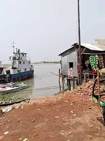 The video shows a rural streetscape, possibly a village or small town in Bangladesh. The environment is characterized by dirt or unpaved roads and basic commercial structures.

People: There are several individuals seen near the shops.

Subjects: The primary subjects are the vehicles known as "easy bikes" or electric rickshaws, which are common in many South Asian countries. These are parked along the roadside.

Objects:
*  Metal or corrugated iron shacks or shops with merchandise visible, including snacks and other small goods.
* A larger body of water is visible in the background. A ship is also visible on the water.
* Electric poles and wires indicate the presence of basic infrastructure.

Actions: The rickshaws are stationary. The video pans and the camera moves along the street.

Interactions: None of the interactions between subjects or objects are clearly displayed in this frame.

Environment:
* The sky is overcast.
* The ground is reddish dirt.
* There are simple structures lining the road.
* The presence of water and boats suggests a river or coastal location.