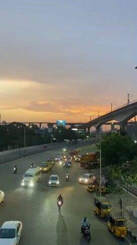 This is a vertical video taken at dusk, showcasing a busy intersection in St. Thomas Mount-Pallavaram Cantonment Board, India. The sky is filled with the warm hues of a sunset, with shades of orange, pink, and grey clouds stretching across the frame. A massive, elevated highway structure, likely for a metro or train line, dominates the upper portion of the scene, casting long shadows over the road below.

The road below is teeming with traffic. Numerous vehicles, including cars, vans, motorcycles, and autorickshaws (tuk-tuks), are moving in both directions. Headlights and taillights create streaks of light, indicating the movement and the low-light conditions. Several autorickshaws are parked on the side of the road. A large yellow truck with a crane is also visible on the road.

People can be seen riding motorcycles and walking on the sidewalk. The overall atmosphere suggests a bustling urban environment during the evening commute. The presence of the elevated railway and the types of vehicles strongly suggest an Indian urban setting. The time of day is clearly evening, as indicated by the sunset and the artificial lighting from the vehicles. There are no discernible words or text visible in the video. The scene conveys the energy and dynamism of a city as it transitions from day to night.