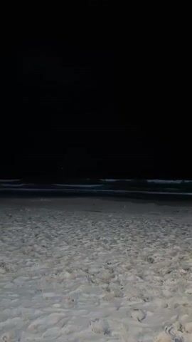 The video shows a nighttime view of a sandy beach with the ocean in the background. The sky is dark and black. Gentle waves are visible breaking on the shore. The sand is light-colored and appears somewhat damp. In the foreground, a white chair with an orange element is partially visible on the right side of the frame. A beam of light, possibly from a flashlight or a spotlight, cuts diagonally across the sky from the upper left to the center of the frame. There are no people clearly visible or interacting in the scene. The overall atmosphere is calm and serene.