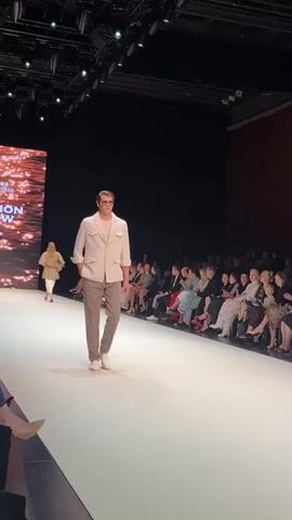 A male model walks down a white runway. He wears a light-colored jacket over a white t-shirt, gray pants, and white slip-on shoes. He also wears sunglasses. He is walking towards the viewer. Behind him, there is a blurred audience seated in rows. To the left of the runway, there is a large screen displaying a sparkling image. The environment appears to be a fashion show.