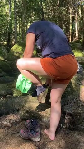 A person wearing a dark blue t-shirt and orange shorts is by a creek in a forest. They are holding a bright green cloth and appear to be washing their foot in the water. Their hiking shoes are on the ground nearby. A black and white dog with a red collar is in the creek, looking towards the person. The environment is rocky and wooded with moss-covered rocks and trees.