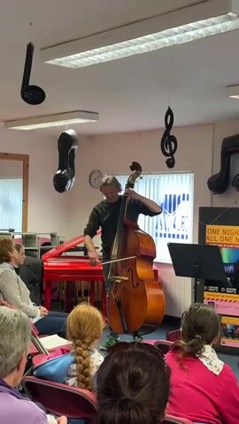 A man plays a double bass in front of an audience. He is wearing a dark green sweater and jeans. The audience consists of at least five people, all facing the musician. The room is decorated with various musical notes, some hanging from the ceiling. A red piano is visible in the background, as well as a window with blinds. There is a music stand in front of the musician and posters on the wall. The lighting suggests an indoor environment, possibly a music studio or a small performance space.