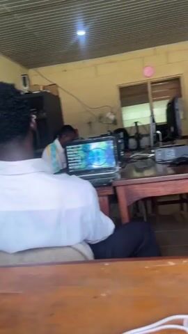 The video shows two young Black men in a room that appears to be a classroom or computer lab. In the foreground, a person wearing a white shirt is seated at a wooden desk, facing away from the camera and looking at a laptop. The laptop screen displays a colorful, abstract image. In the background, another person wearing a gray jacket and a red and white beanie is seated at a similar wooden desk, holding a smartphone and looking at it. There are several computer monitors, a projector, and wires on the desks. The walls are painted a light yellow, and the ceiling has a corrugated metal finish. There are windows with blinds and a door visible in the background. A round pink clock is mounted on the wall. The lighting in the room is somewhat dim.