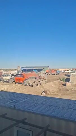 A bright blue sky fills the top half of the vertical frame. Below the sky, an open, dusty lot is visible, populated by various trucks and equipment. In the foreground, a section of a corrugated metal roof with a window and some wooden beams is visible. The lot appears to be a construction or industrial site. In the middle ground, several trucks are parked, including a prominent orange tanker truck with a dark-colored trailer. Further back, more vehicles and structures, possibly storage containers and a building with a green roof, can be seen. The ground is uneven and appears to be composed of dirt and gravel. The overall impression is of a working yard under a clear, sunny sky.