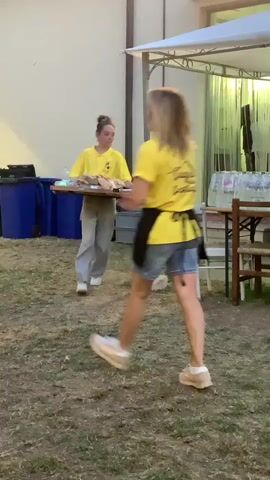 A young woman in a yellow t-shirt and denim shorts, with a black apron, walks from left to right across a grassy area. She has blonde hair and is wearing white sneakers. She is carrying a tray with a red bin bag behind her on the left. In the background, a young woman in a yellow t-shirt and grey pants is walking from left to right, carrying a wooden tray with items on it. She has her hair in a bun and is wearing white sneakers. Behind her, there is a white tent structure, tables with chairs, and bottles of water. The environment appears to be an outdoor event or gathering.