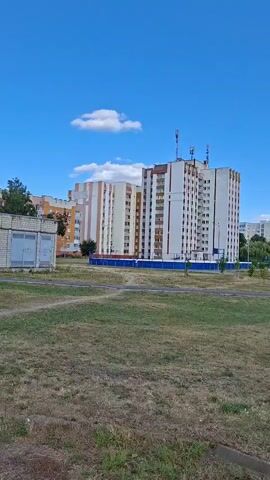 The video pans across an outdoor scene on a sunny day with a bright blue sky and scattered white clouds. In the foreground, there is a patch of dry, mowed grass with some greener patches and a few small stones. In the middle ground, there is a paved pathway or road. Beyond the pathway, there is a large, multi-story apartment building with white and beige facades, featuring many windows and balconies. The building has several antennas on its roof. To the right of this building, there are several tall pine trees. Further to the right and in the background, more apartment buildings and trees are visible. The camera continues to pan, revealing a lower, brick building with white window frames. The overall environment appears to be a residential area or a park adjacent to residential buildings. There are no people or animals visible in the video.
