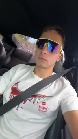 A young male with short brown hair wearing sunglasses and a white t-shirt with a red graphic logo is sitting in the driver's seat of a white car, looking to his right. The car is moving along a road with a rocky, wooded hillside to the right. The interior of the car shows black leather seats and a steering wheel. The camera is positioned outside the car, looking in through the open driver's side window.