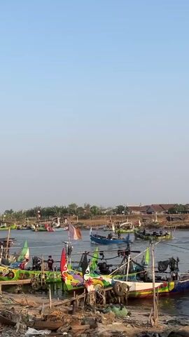 The video shows a body of water with many colorful fishing boats. In the foreground, several boats are docked along a shore that appears to be cluttered with debris. Some of these boats are brightly painted with intricate designs. In the background, more boats are on the water, and some appear to be moving. There is a clear blue sky above, and a hazy horizon with trees and buildings on the shore. The scene depicts a busy fishing harbor.