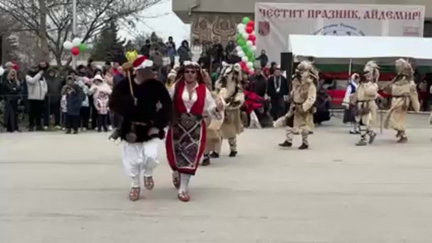 The video shows a public event in what appears to be a town square or public space. A small crowd of people, including adults and children, stand watching a procession. Several individuals dressed in traditional Bulgarian costumes are dancing, and there are also people in sheepskin costumes wearing ornate masks. One individual is wearing a Santa hat over their traditional attire. In the background, there's a building with a banner that says "Честит Празник, Айдемй." There are also balloons in the colors of the Bulgarian flag. The weather appears to be overcast and cold.
