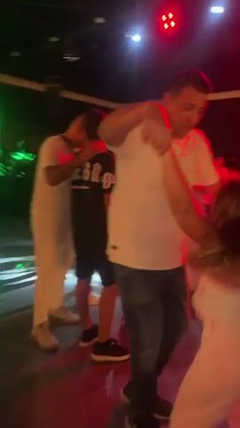 This video shows a crowded nightclub or party scene with people dancing under colorful lights. In the foreground, a man in a white t-shirt and jeans is dancing with a woman wearing a white strapless top and light-colored pants. To the left, a young person in a black t-shirt and shorts is also dancing. Other people are visible in the background, dancing and socializing. The lighting is dynamic, with flashes of purple and green.