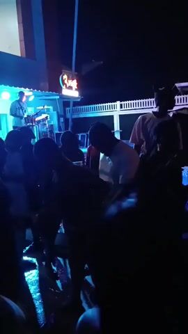 The video shows a crowd of people gathered at night to watch a live music performance. A DJ is visible on a small stage set up outside a building with a sign that says "Ritz." The stage is lit with blue and white lights. The crowd is mostly silhouetted against the light, making it difficult to discern individual features. Some people are moving, possibly dancing to the music. The location appears to be an outdoor area near a building, with a white fence visible in the background.