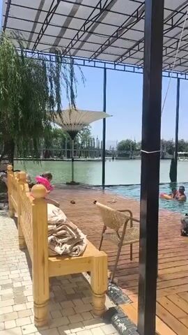 The video shows a resort with a pool and a lake. People are swimming in the pool and some are relaxing on lounge chairs. There are inflatable toys in the pool, including a crocodile. The weather appears to be sunny and warm.