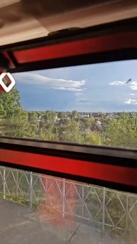 The video pans across a view from a window, looking out over a village. The window frame is red and black. In the foreground, there is a balcony with a metal railing and a concrete floor. Beyond the balcony, there are many trees and houses with red roofs. The sky is partly cloudy with some patches of blue. There are some objects that appear to be bugs flying in front of the window.