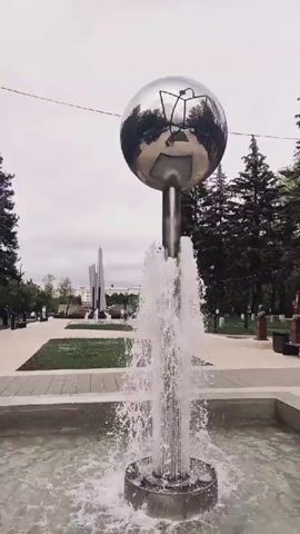 The video shows a fountain in a park. A large, reflective spherical sculpture with an abstract drawing on it sits atop the fountain's central column. Water sprays upwards from the fountain's base, creating a misty effect around the sculpture. The park appears to be paved with light-colored stones, and there are trees and some other sculptures in the background. The sky is overcast. In the reflection on the sphere, two people are visible walking on the path.