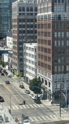 The video shows an elevated view of a city street on a sunny day. Tall buildings line both sides of the street. Cars are driving and parked along the street. Pedestrians are visible on the sidewalks and crossing the street. One person is riding a bicycle across the intersection. Traffic appears to be moderate. The buildings are a mix of architectural styles, some appearing to be older and others more modern. Construction is visible on the lower right side of the frame.