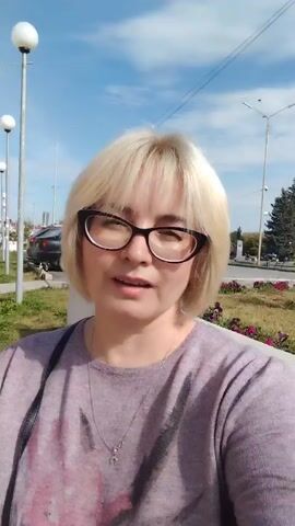 A woman with blonde shoulder-length hair, wearing glasses and a light purple shirt with a floral pattern, smiles at the camera. She is outdoors on a sunny day, with a building in the background and a street with a lamppost. The video appears to be a selfie.