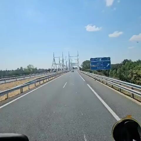 A first-person perspective video shows a vehicle driving across a bridge on a sunny day. The road is asphalt with white lane markings. On the right side of the road, a blue highway sign indicates destinations: Sahagun 500m, Carrion de los Condes, and Palencia. The bridge has a distinctive white truss structure overhead and yellow guardrails. Trees and vegetation line the sides of the road. The sky is clear blue with a few scattered white clouds. There are other vehicles on the bridge, and the overall impression is of smooth travel on a well-maintained road.