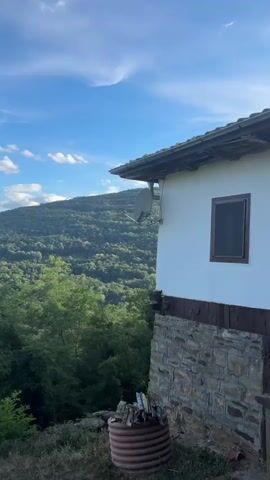 The video shows a panoramic view of a lush, green mountainous landscape under a bright blue sky dotted with white clouds. In the foreground, a rustic white building with a stone foundation and a brown tiled roof is partially visible on the right side of the frame. A satellite dish is mounted on the side of the building. In front of the building, a small pile of firewood is arranged in a cylindrical metal holder. The camera pans across the expansive valleys and rolling hills covered in dense forestation, revealing layers of mountains receding into the distance. The overall impression is one of a serene, natural setting viewed from an elevated vantage point.
