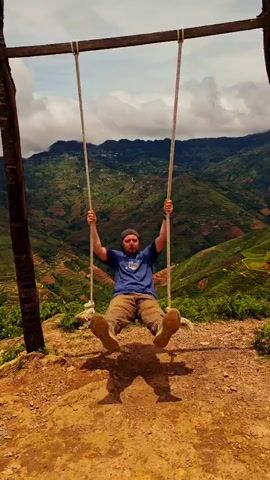 A man sits on a wooden swing suspended by ropes from a wooden frame. The swing is positioned on a dirt surface overlooking a vast landscape of green terraced hills under a cloudy sky. The man is wearing a blue t-shirt, brown pants, and a cap. He is holding onto the ropes of the swing and appears to be moving back and forth. The video shows the man from a static frontal view, with the swing and the background landscape filling the frame.