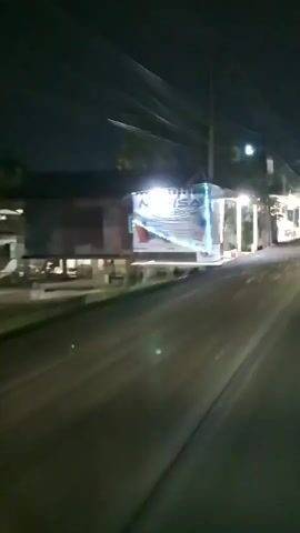 The video shows a nighttime view from a moving vehicle. The vehicle is traveling on a paved road, and the camera is pointed towards the right side of the road. Along the side of the road, there are several small structures that appear to be shops or market stalls. These structures are illuminated by artificial lights, and some have signs and decorations, including what looks like string lights. There are also utility poles and power lines visible overhead. The overall impression is of a small commercial area at night.