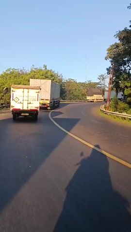 A motorcycle with two riders is traveling on a paved road in a rural area. The riders are casting a shadow on the road. There are several trucks on the road ahead. The road is curved and lined with trees and vegetation. The sky is clear and blue. The sun appears to be setting, casting a warm light on the scene.