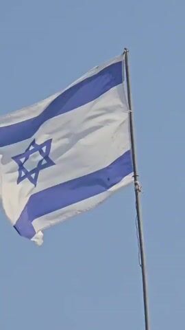 A close-up shot focuses on the Israeli flag as it waves gently in the wind against a clear blue sky. The flag has a white background with two horizontal blue stripes near the top and bottom edges. In the center, a blue Star of David is prominently displayed. The flagpole is visible on the right side of the frame, supporting the flag. The movement of the flag suggests a mild breeze. The sky is cloudless and a uniform pale blue.