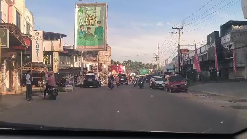 The video shows a street scene in what appears to be a town or city in Indonesia. It is daytime with a clear sky. The camera is mounted inside a vehicle driving down the street. There are buildings on either side of the road, many appearing to be shops or businesses. There are cars, motorcycles, and a few pedestrians on the street. A large billboard advertising what appears to be a political campaign is visible above the buildings on the left. There are power lines overhead. The traffic appears to be light. The video conveys a sense of everyday life in a busy commercial area.