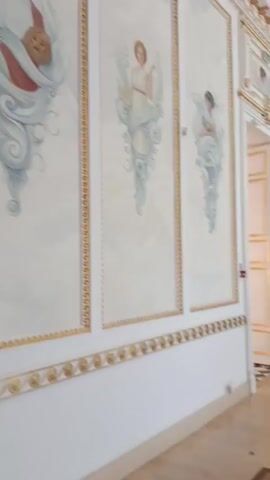 The video pans across a grand hall, showcasing ornate white walls with gilded decorative molding along the ceiling and borders of the painted panels. Two tall, narrow painted panels are visible on the left wall, each featuring a figure in classical attire amidst swirling blue and white drapery. The panel on the left depicts a person holding a mask and a dagger, while the panel on the right shows a figure holding an open book. The floor is a polished wood with a geometric inlay pattern. Further down the hall, an open doorway reveals another room. A large, ornate fireplace with red marble and gilded details stands against the wall, accompanied by a black speaker on a stand. A gilded mirror hangs above the fireplace. Multiple large, multi-tiered chandeliers with many lights hang from the high, decorated ceiling. Several people are scattered throughout the hall, some looking at the paintings and architectural features, while others appear to be conversing. The overall impression is one of opulent historical architecture.