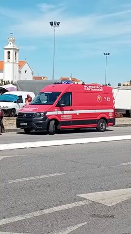 Here is a description of the video.
The video shows a red ambulance parked on the side of a road. The ambulance has the words "BOMBEIROS CANTANHEDE" written on the side and features the emergency number, 112. There are several cars parked nearby, and a few people are standing near the ambulance. In the background, there is a white church with a tall steeple. The sky is blue and the weather appears to be sunny. The scene looks like it could be in a town square or a similar public area.