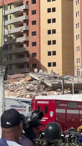 The video shows the aftermath of a building collapse. In the foreground, the backs of several people's heads are visible, including one person wearing a black baseball cap and another wearing a black motorcycle helmet. To the right of them, a red emergency vehicle with "EMERGENCIAS" and "UNIDAD" written on its side is parked. To the left of the red vehicle is a white ambulance with "AMBULANCIA" and "EMERGENCIA" written on it. In the background, a large pile of rubble from a collapsed structure is visible. Behind the rubble, portions of two multi-story buildings stand intact. One building has reddish-brown brick facade and the other has a tan facade. Satellite dishes are visible on the debris.
