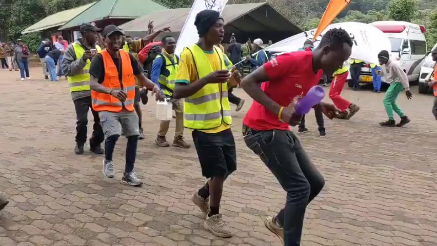 The video shows a group of approximately 15 people dancing outdoors in a circle. Most of the people are wearing light-colored reflective vests, some are wearing backpacks or carrying objects like plastic containers or metal jerrycans. Several are holding or positioned near triangular flags with words and logos.  Behind the dancers are several vehicles including an ambulance and a minibus.  There is a building with a metal roof in the background, and a tree-covered hill in the far distance. The ground is paved with interlocking bricks. The sky is overcast.  The people are dancing energetically, moving in a counterclockwise direction.