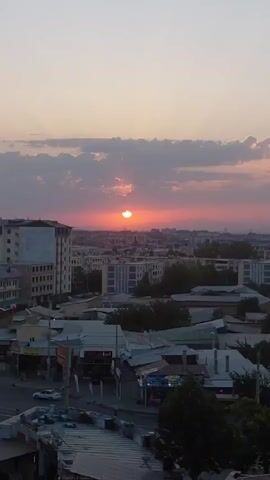 This is a vertical, high-angle shot of a cityscape at sunset. The sun, partially obscured by clouds, is a vibrant orange disc near the center of the frame, casting a warm glow across the sky and the city below. The sky transitions from a pale pink and orange near the horizon to a light grey and blue at the top of the frame. Puffy, grey clouds are scattered across the sky, some catching the sunset's hues.

The scene depicts a dense urban environment with a mix of modern and older architecture. In the foreground, the rooftops of various buildings are visible, some with corrugated metal coverings. A few trees are interspersed among the buildings. Further back, taller apartment buildings with multiple windows stand out, one a light beige color and another with blue accents. The cityscape extends into the distance, with more buildings and faint outlines of mountains on the horizon.

At street level, signs of commercial activity are evident. In the lower-left quadrant, a white car is parked on the street. Several shops and businesses are visible, their storefronts facing the street. One sign clearly reads "MINTAY" in white lettering on a blue background. Another sign displays text that is difficult to read but appears to be in a Latin-based alphabet.

The overall impression is a calm and picturesque evening scene. The time of day is clearly sunset, indicated by the sun's position and the warm colors in the sky. The presence of clouds suggests a partly cloudy sky. The urban setting, combined with the specific signage, points to a location in Samarkand City, Uzbekistan. There is no visible human activity or specific event taking place, with the focus being on the natural beauty of the sunset over the city.