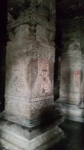 The video shows the interior of a dimly lit, ancient stone structure, likely a temple or cave. Several large, intricately carved stone pillars line the space. People are walking through the structure, some in pairs and others individually. One man in a blue t-shirt and jeans walks away from the camera, followed by a woman in a patterned sari. Another woman, wearing a maroon top and carrying a backpack, walks in the same direction. The floor is wet and reflective, suggesting it might be damp or recently cleaned. Lighting appears to be minimal, with some artificial lights casting pools of illumination on the floor and highlighting the carvings on the pillars. The atmosphere is quiet and reverent, consistent with a historical site.