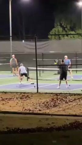 Four individuals are engaged in a doubles pickleball match on an outdoor court at night. The court is well-lit by overhead lights. The players are dressed in casual athletic wear, with two wearing hats. The court is surrounded by a chain-link fence, and beyond the fence is a patch of grass covered in fallen leaves. The camera angle is slightly elevated, positioned outside the court perimeter. The game is in progress, with players moving around the court and hitting the pickleball.